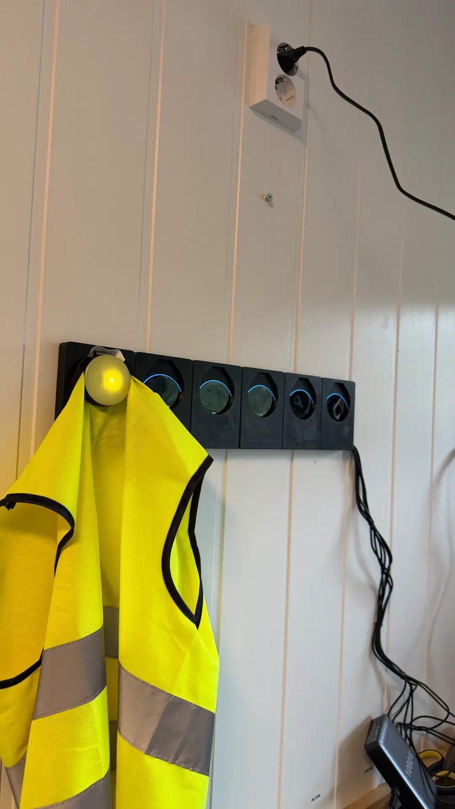 Beehive-sensoren festet på en gul refleksvest, ved siden av ladestasjonen i barnehagens garderobe